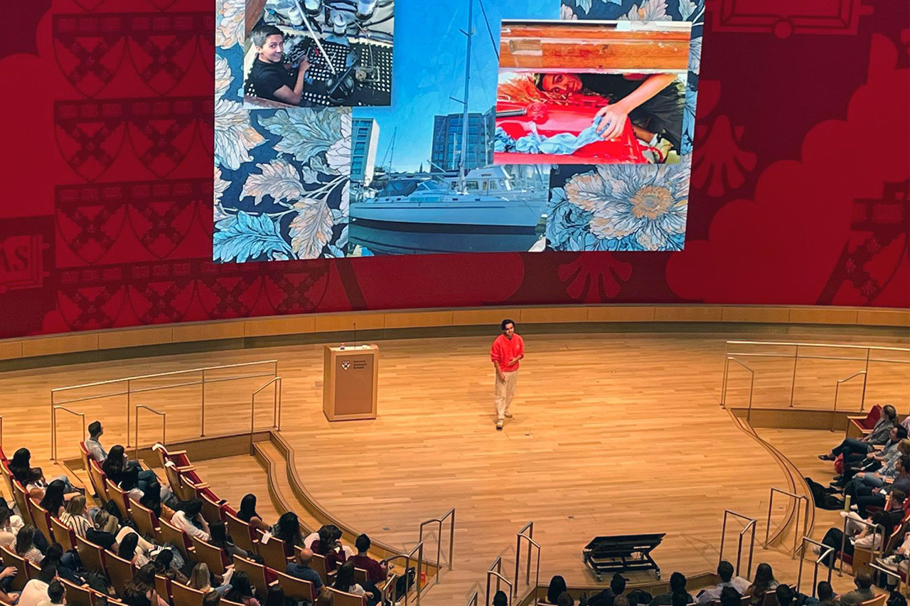 A white man in a red sweater is speaking on a Harvard Business School stage to a large audience. Behind him is a large screen showing a collage of images of him on a boat as a child.