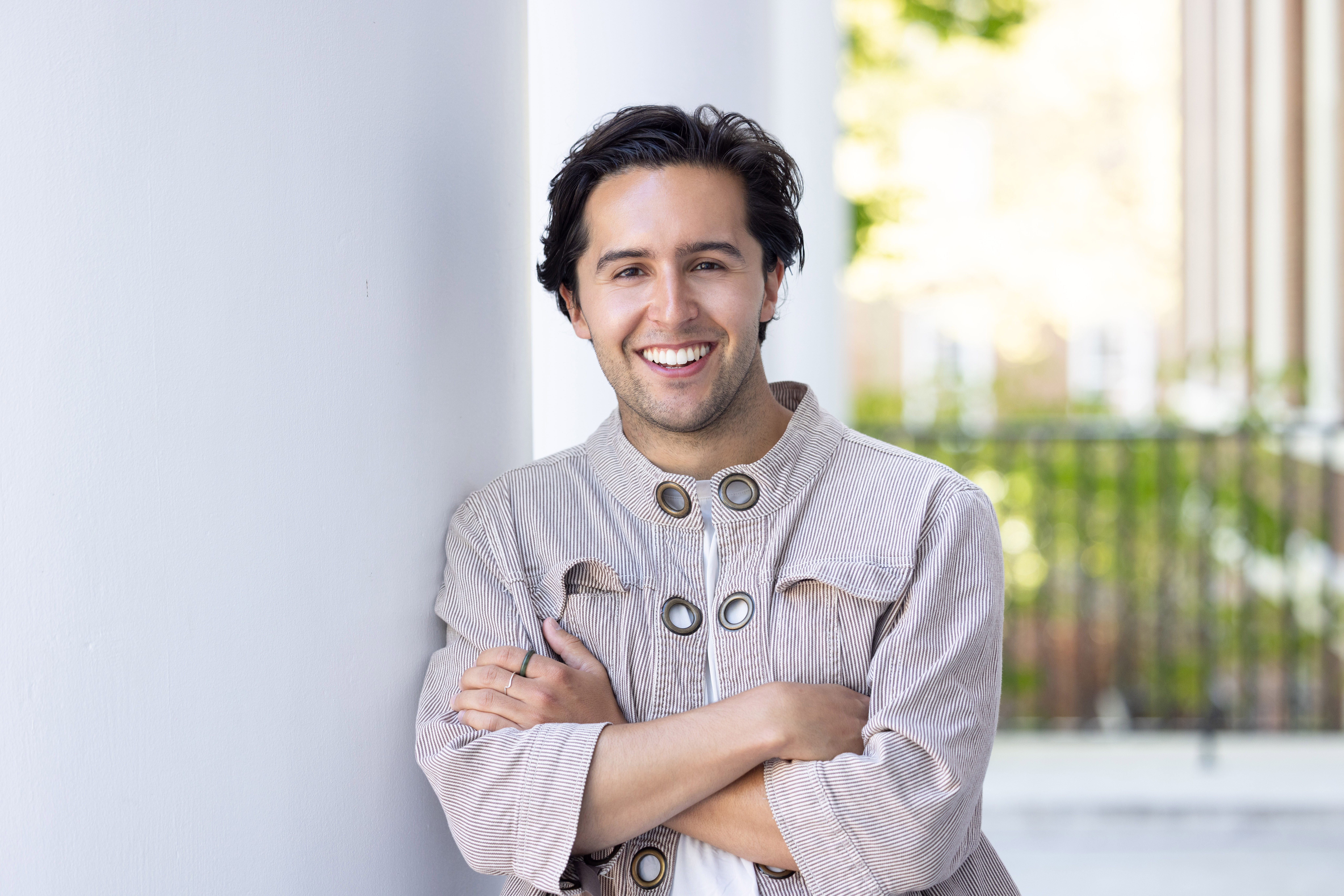 Smiling person with dark hair in a light striped jacket leans against a white pillar outdoors.